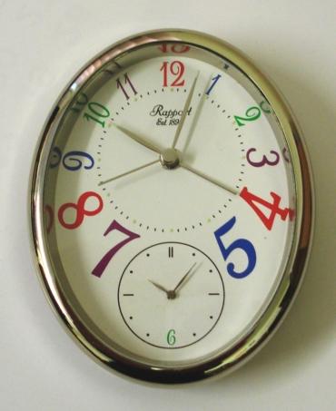 White metal  dual time quartz alarm clock by the Rapport Company. White painted dial with silver coloured hands in 2 time displays. Top display includes the alarm setting hand and a sweep seconds. The back of the clock has alarm on/off and snooze buttons, and a fold away support strut.