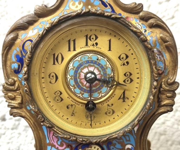 French rococo style 8 day time piece clock in a heavily decorated enamelled gilded brass case circa 1900, made in Paris and retailed by Brook & Son, Edinburgh. Ornate gilt brass bezel with chamfered flat glass over a circular gilt dial with black Gothic hours and matching pierced steel hands with central enamelled floral style disk. Plain French key wound brass drum movement numbered 542 with replacement jewelled lever escapement platform. Height - 9" Width - 4.5" Depth - 2.5".