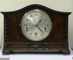 8 day dark stained oak cased Westminster chime mantel clock circa 1920. Oblong case with wave top and integral barley twist columns and applied decorative mouldings. Circular brass bezel with convex glass over a silvered dial with black arabic hours and blued steel hands. Square brass spring driven, rod striking movement with decorative wriggle work back plate, stamped #2113, and a vertically mounted French lever escapement.  Dimensions: Height - 10", width - 13.75", depth - 8.25".