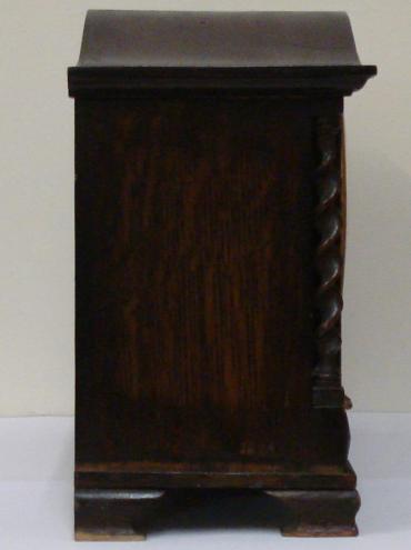8 day dark stained oak cased Westminster chime mantel clock circa 1920. Oblong case with wave top and integral barley twist columns and applied decorative mouldings. Circular brass bezel with convex glass over a silvered dial with black arabic hours and blued steel hands. Square brass spring driven, rod striking movement with decorative wriggle work back plate, stamped #2113, and a vertically mounted French lever escapement.