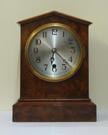 American 8 day burr walnut veneer cased mantel clock timepiece circa 1900. Simple arch topped case with integral plinth. Gilt brass bezel with flat glass over a silvered dial with black arabic hours and black steel moon hands. Simple brass, spring driven pendulum regulated, movement which because of the solid casework construction, only accessible from under the clock.