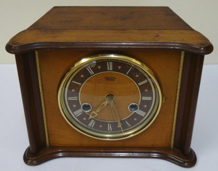 English Smiths Enfield 8 day wood veneer cased gong strike mantel clock circa 1950. Unusually shaped flat top case with gilt bezel and domed glass over a dark brown chapter ring with white roman hours and pierced gilt hands. Rear door to brass spring driven, floating balance movement with slow / fast regulation. Time adjust and winding squares behind the front face glass.