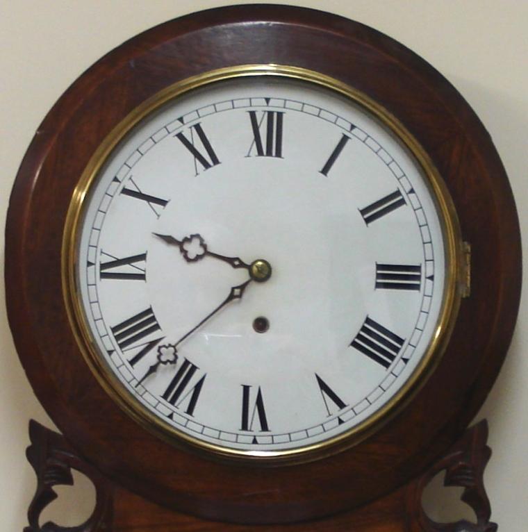American Ansonia Clock Co. round faced extended drop dial 8 day wall clock in a carved and moulded mahogany case with scroll bottom. Brass bezel with flat glass over a white painted dial with black roman numerals and ornate black painted hands with separate lower in view brass pendulum window. Spring driven, pendulum regulated, time piece movement, circa 1890.  Dimensions: Height - 31", Width - 16.5", Depth 4.5".