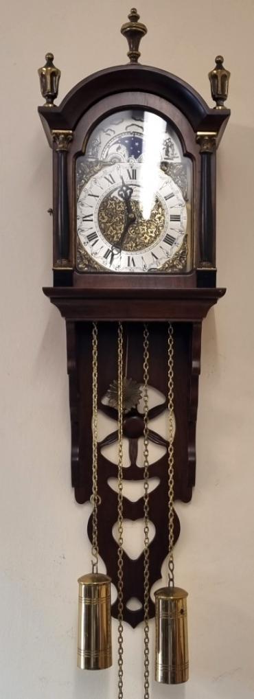 Modern Reproduction Dutch style wall clock by St James of London. Mahogany hooded top case with brass finials and opening front with brass mounted side columns and fret worked side panels and lower bracket base. Flat glass front over highly decorated brass dial plate with silvered chapter ring and black Roman hours and hands together with applied angel spandrels all surmounted by a lunar calendar and moon phase indicating panel. German 8 day weight driven, pendulum regulated movement striking the half hour and hour on a bell, with decorative sun burst pendulum and turned polished brass weights. Case Dimensions: Height - 27", Width - 8", Depth - 6".