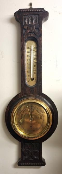 English carved dark oak mounted aneroid barometer and thermometer, circa 1920s. Circular brass bezel with flat glass over a polished brass dial engraved with black inches of mercury pressure index. Black pressure indicating hand with gilt manual history marker together with a brass mounted thermometer displaying temperature in both Fahrenheit and Centigrade. Height - 18" and width - 6.5".