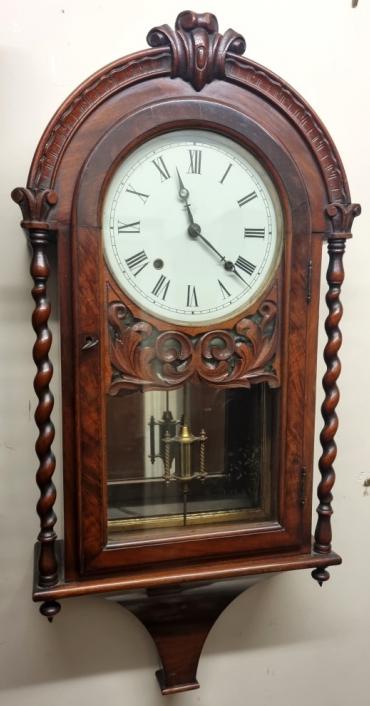 American mahogany cased 8 day striking wall clock. Very decorative carved and moulded mahogany case with barley twist columns, round top and opening glass front door. White dial with black Roman hours and blued steel hands and the two winding squares, together with lower mirror backed brass pendulum. American 8 day spring driven, pendulum regulated movement striking on the polished silvered bell. Case Dimensions: Height - 33", Width - 15", Depth - 5.5".